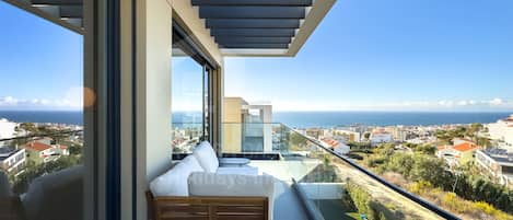 A balcony with a wooden bench, a glass balustrade, and a view of the sea and town.