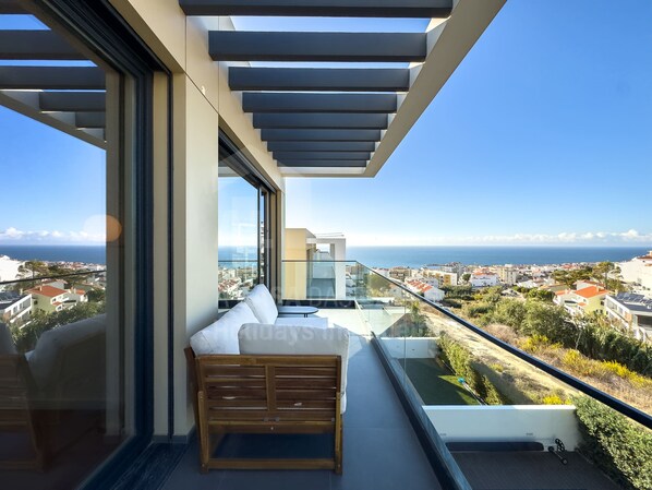 Un balcón con banco de madera, barandilla de cristal y vista al mar y a la ciudad.