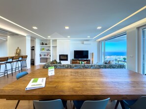 Una sala de estar moderna con una gran mesa de madera, un sofá y un televisor. Se ve el océano a través de amplias ventanas.
