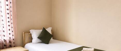 A single bed with a wooden frame, a nightstand with drawers, a green throw blanket, and a patterned carpet.