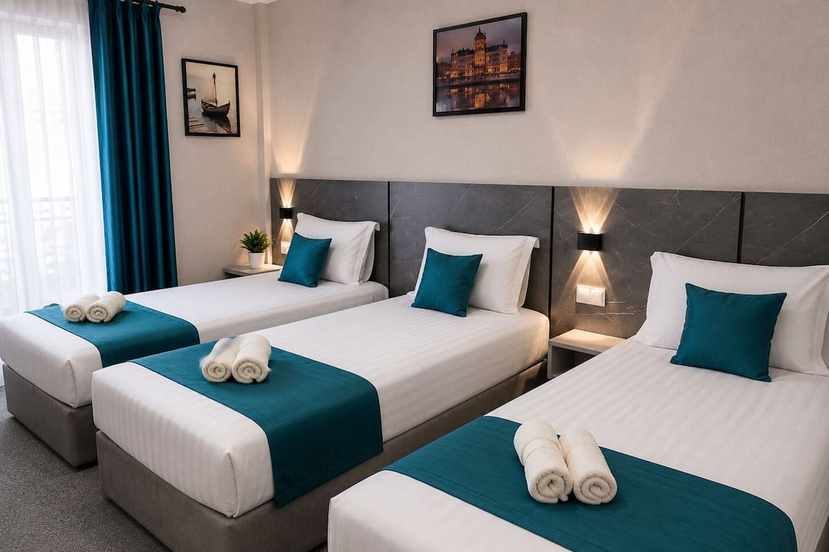 Une chambre d’hôtel avec trois lits simples, chacun recouvert d’une couvre-lit turquoise et agrémenté d’oreillers blancs, une table de chevet avec une lampe et un tableau encadré au mur.