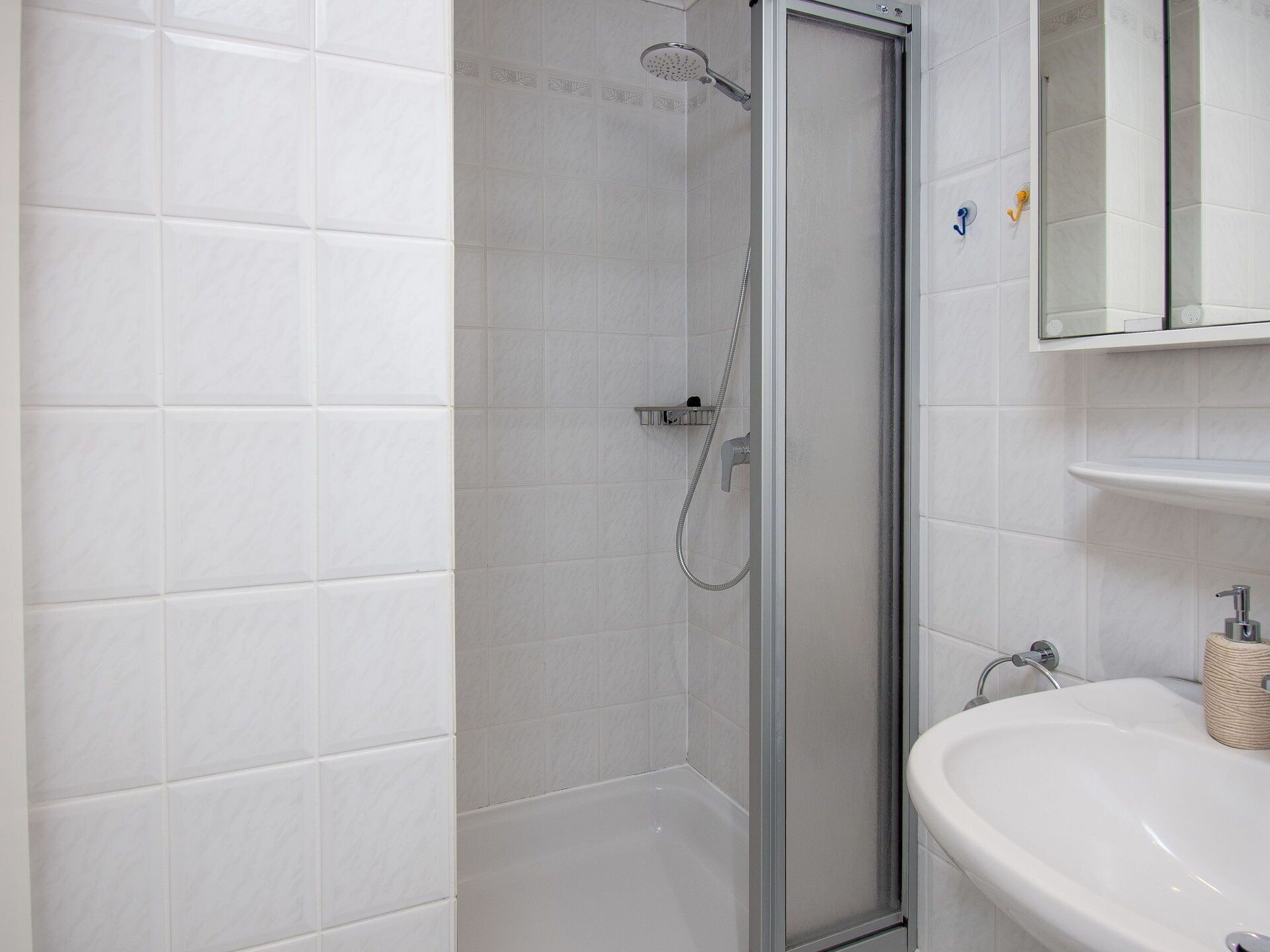Une salle de bain moderne avec des carreaux blancs, une douche en verre et un lavabo blanc doté d’un distributeur de savon.