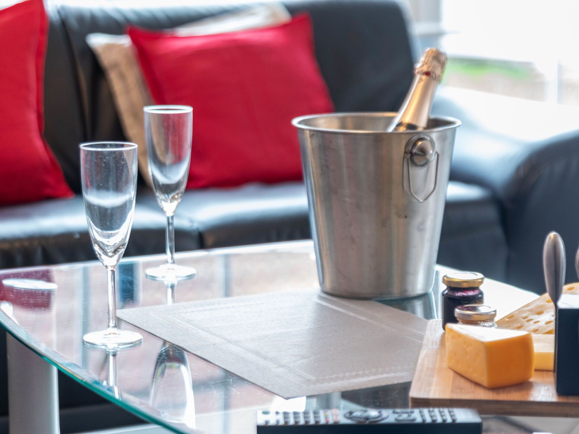 Ein Glastisch mit zwei Sektgläsern, einem Metallkübel mit einer Champagnerflasche und einem Holzbrett mit Käse und Beilagen.