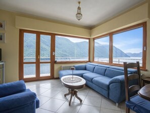 Ampla sala de estar com uma grande janela com vista para um lago e montanhas, um sofá azul, uma pequena mesa redonda e um candelabro.