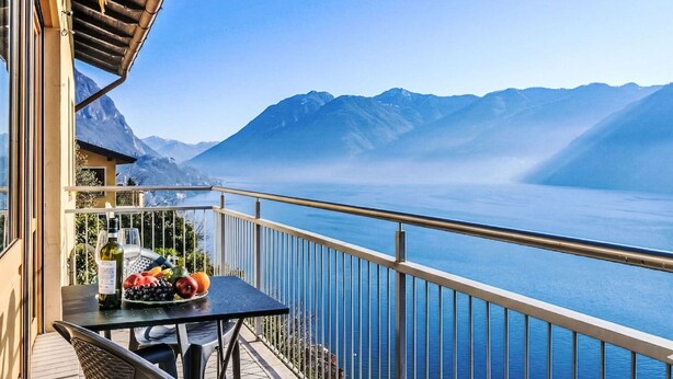 Ein Balkon mit Tisch, gedeckt mit Obst und Wein, mit Blick auf einen See und Berge im Hintergrund.