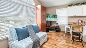 A living room with a sofa, TV, wooden cabinets, and a kitchen area with a bar stool. - Sunshine Hideaway - Team Joseph Ellen (Jacksonville Beach)