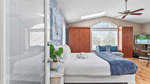 A bedroom with a bed, a ceiling fan, a TV, and a window with blinds. - Sunshine Hideaway - Team Joseph Ellen (Jacksonville Beach)