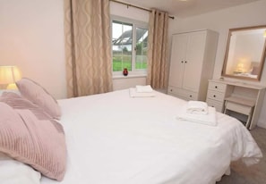 A neatly made bed with pillows, a bedside lamp, a window with curtains, a wardrobe, and a mirror.