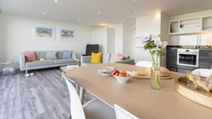Un salon moderne avec un canapé gris, une table à manger ornée de fruits et d’un vase, et une cuisine avec des meubles blancs et des appareils électroménagers.