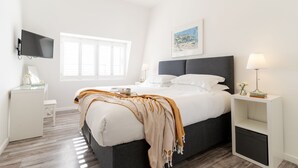 A modern bedroom with a bed, bedside tables, a TV, and a window with blinds. - 7 Beachview - apartment in Crantock sleeps 4 guests in 2 bedrooms (Crantock)