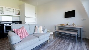A modern living room with a grey sofa, a small round table, and a kitchen area with white cabinets. - 8 Beachview -  apartment in Crantock sleeps 4 guests in 1 bedroom (Crantock)