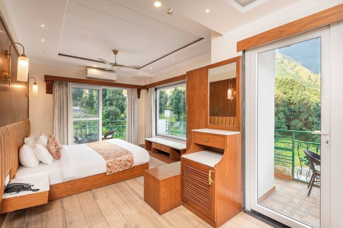 A bedroom with a bed, bedside table, and a large window offering a view of greenery.