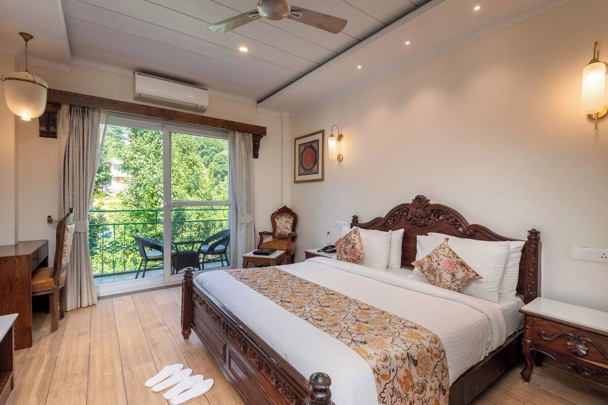 A bedroom with a large bed, a desk, a chair, and a balcony with a view of greenery.