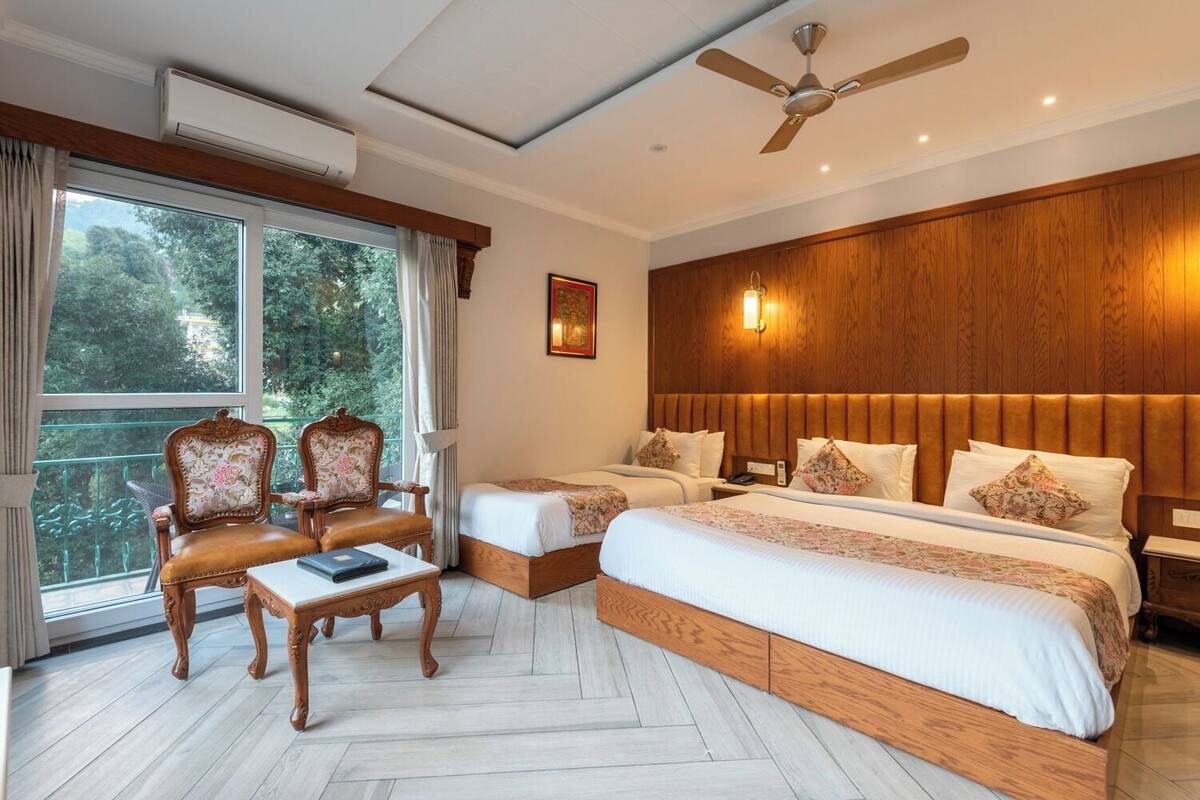A hotel room with a large bed, two smaller beds, a wooden headboard, a desk, two chairs, a ceiling fan, and a large window with curtains.