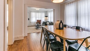 A dining area with a wooden table and chairs, a chandelier, and a view into a living room with a sofa and curtains.