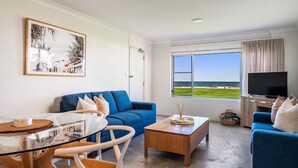 A living room with a blue sofa, a glass coffee table, a TV, and a large window with a view of the sea. - Absolute Beachfront Apartment 3B (Lennox Head)