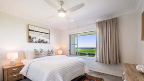 A bedroom with a bed, nightstands, a ceiling fan, a window with curtains, and a view of a green landscape. - Absolute Beachfront Apartment 3B (Lennox Head)