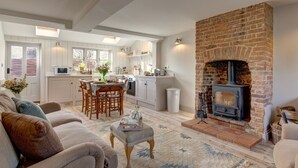 A cozy living room with a fireplace, a dining area, and a kitchen with white cabinetry and a brick accent wall. - The Flints - sleeps 3 guests  in 2 bedrooms (Langham)