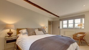 A bedroom with a large bed, bedside lamps, a wicker chair, and a window with curtains. - The Flints - sleeps 3 guests  in 2 bedrooms (Langham)