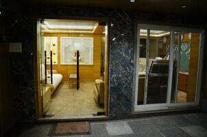 Entrada de hotel com balcão de recepção, área de estar com sofá e cadeiras, e banheiro visível.