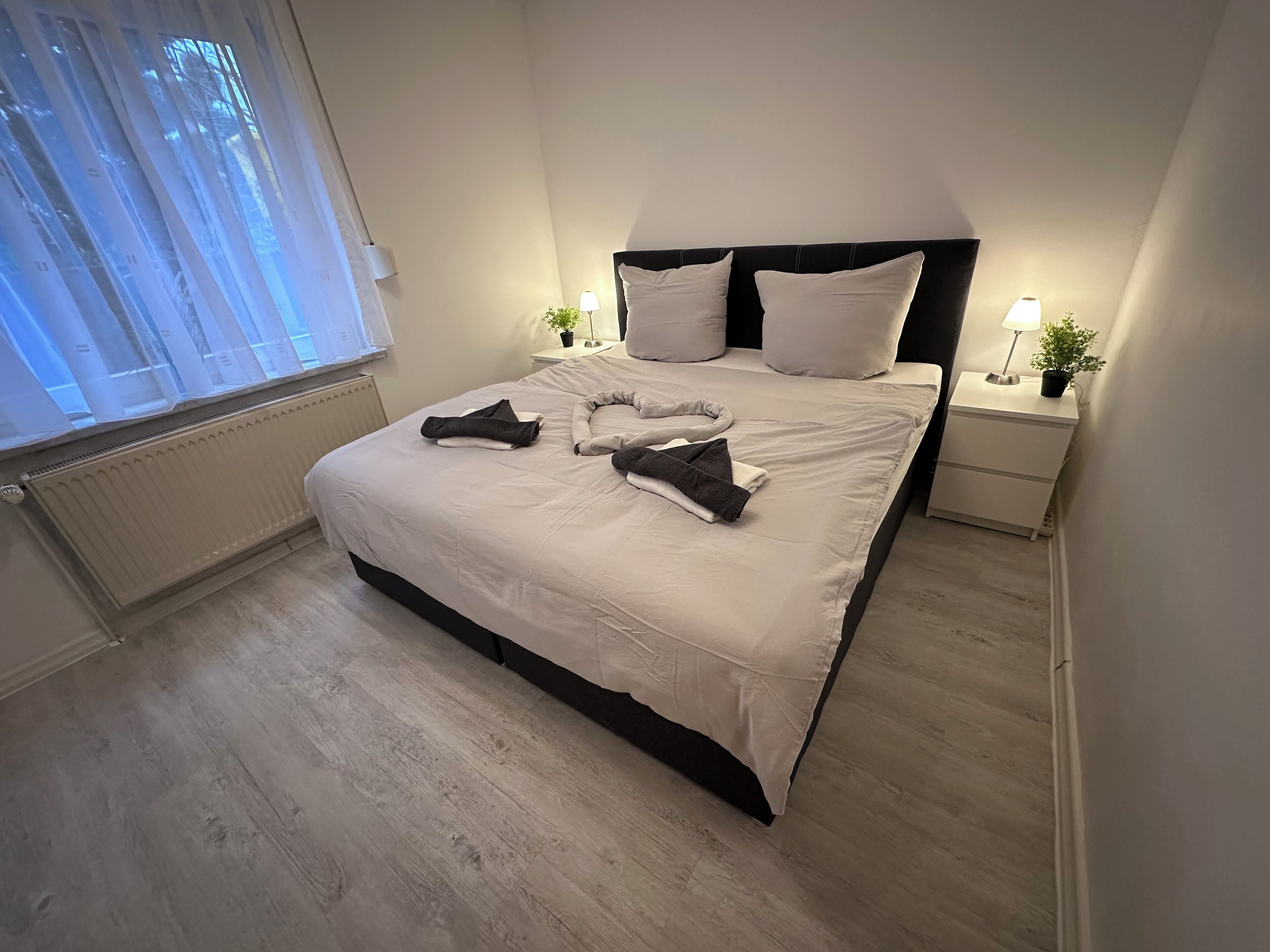 Un lit bien fait, avec des oreillers et des serviettes pliées, une table de chevet avec une lampe et une petite plante, et une fenêtre ornée de rideaux légers.