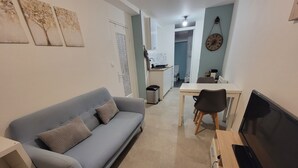 En moderne stue med en grå sofa, et spisebord med stole, et fjernsyn og et køkken i baggrunden.