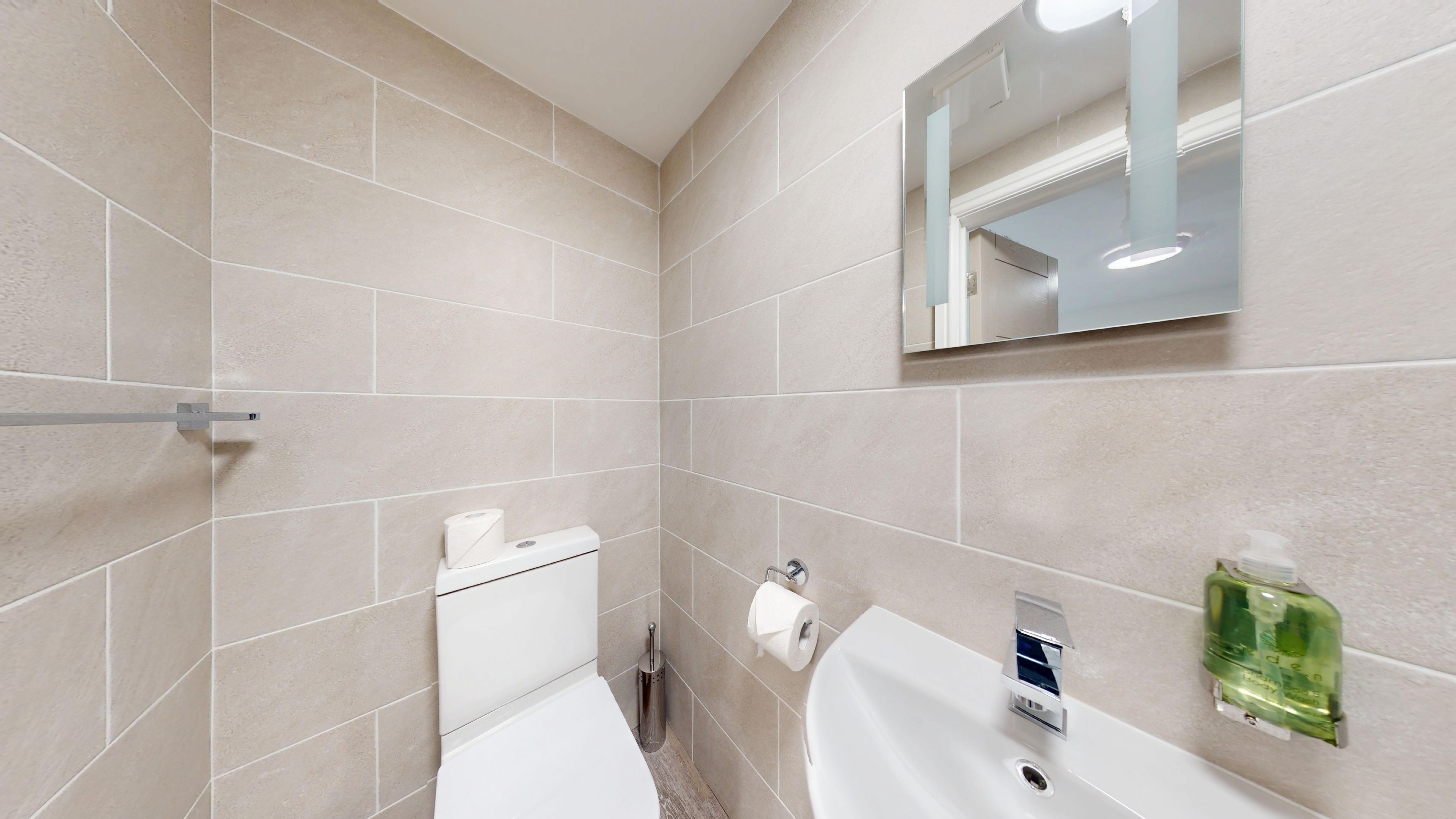 A bathroom with beige tiles, a toilet, a sink, a mirror, and a hand soap dispenser.