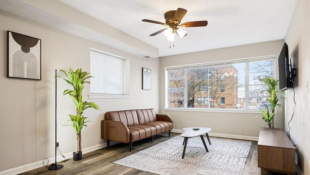 A living room with a brown leather sofa, a white coffee table, a patterned rug, a ceiling fan, and a large window with a view of buildings outside. - A Trendy Escape close to Downtown Peoria (Peoria)