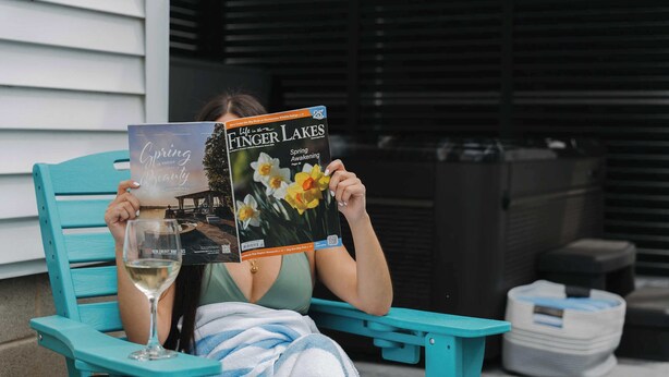 Una persona seduta su una sedia color turchese, che tiene in mano e legge un libro intitolato 'Life on the Finger Lakes'.