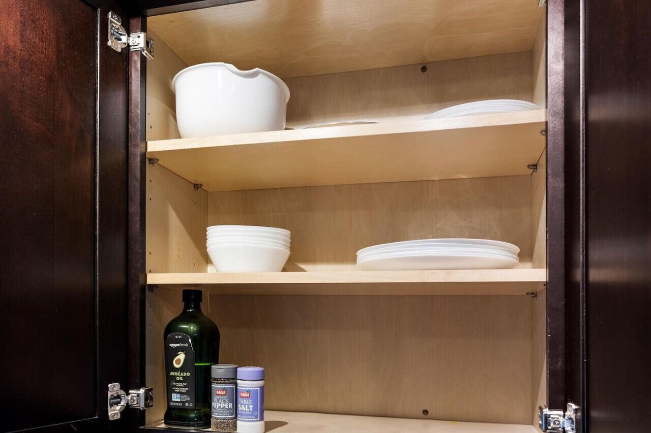 A wooden cabinet with three shelves, each holding white bowls and plates. Below the top shelf, there are bottles of olive oil and table salt.