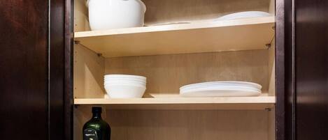 A wooden cabinet with three shelves, each holding white bowls and plates. Below the top shelf, there are bottles of olive oil and table salt.