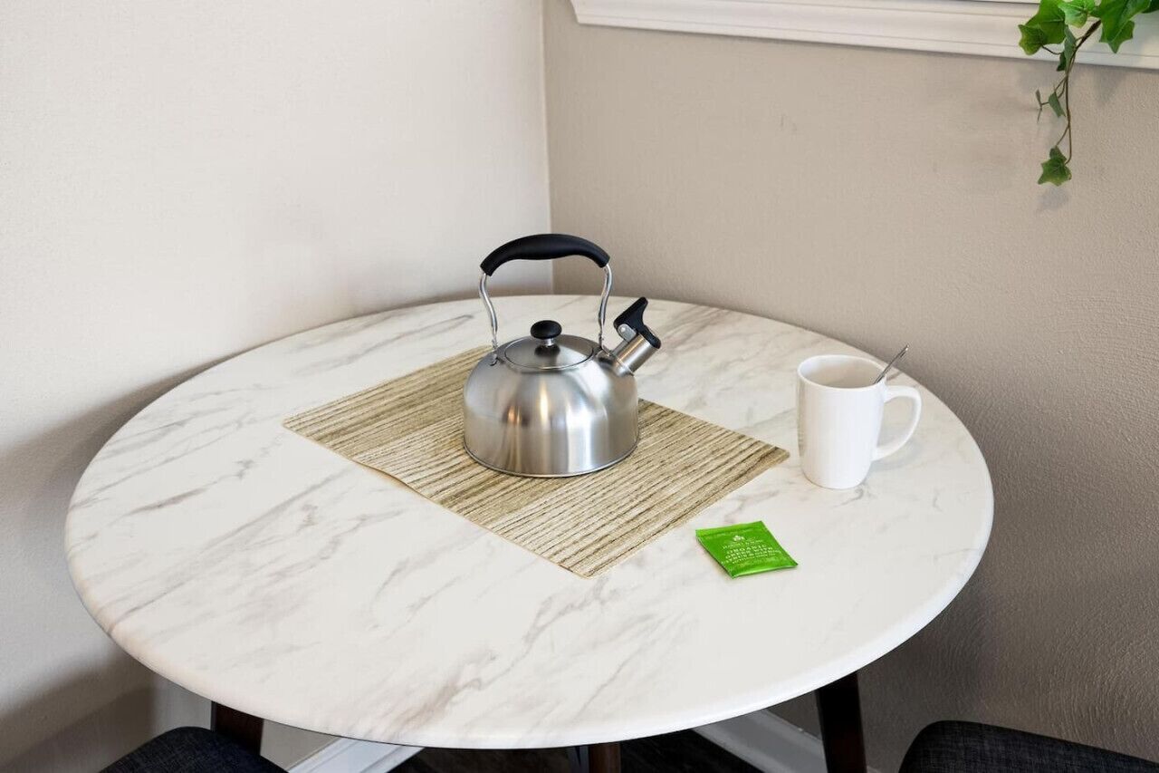 Ein runder Tisch mit weißer Marmorplatte, einem Edelstahl-Wasserkocher, einer weißen Tasse und einer grünen Packung auf einer gewebten Tischmatte.