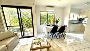 A modern living room with a sofa, a coffee table, and a dining area with chairs and a table. There is a kitchen area with cabinets and a sink.