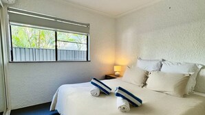 A hotel room with a bed, a ceiling fan, a window with a view of greenery, and a nightstand with a lamp.