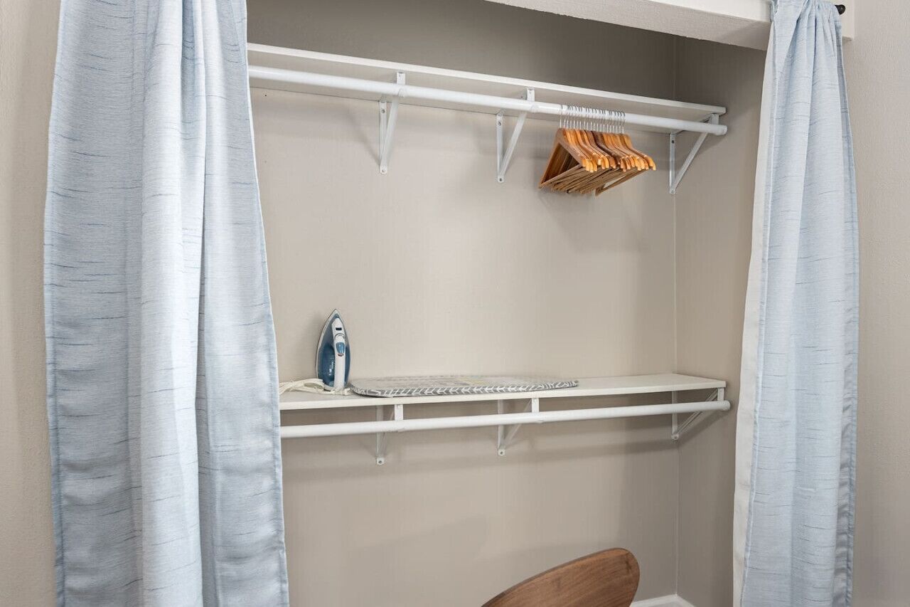 Un petit espace clos comprenant une étagère blanche, un portant avec des cintres en bois, et un seul fer à repasser posé sur l’étagère.