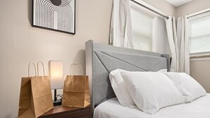 Habitación de hotel con cama, mesita de noche con dos bolsas de papel marrón, lámpara e imagen en un marco en la pared.