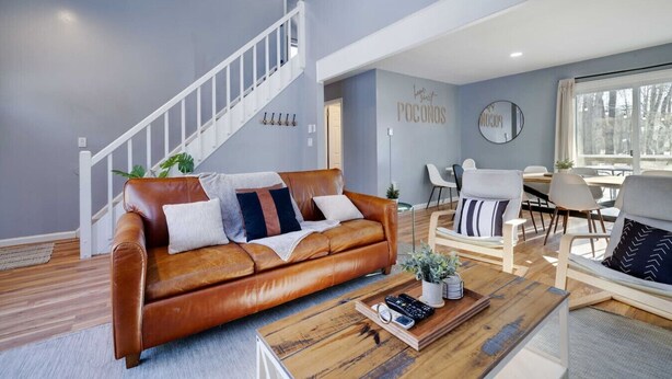 A modern living room with a brown leather sofa, a wooden coffee table, and a dining area with white chairs. - Pocono Lake Charmer: Lakeview Retreat for 10 (Tobyhanna)