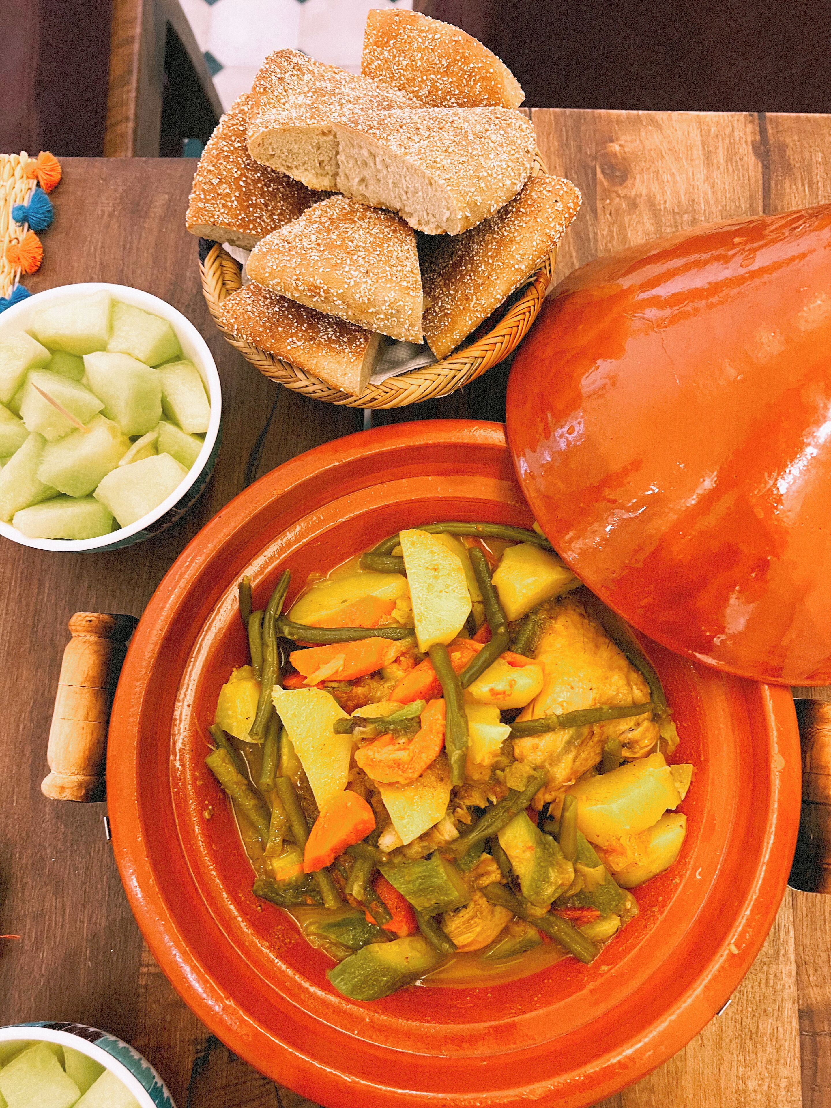 A traditional Moroccan tagine with vegetables and meat, a bowl of cubed melon, and a basket of bread.