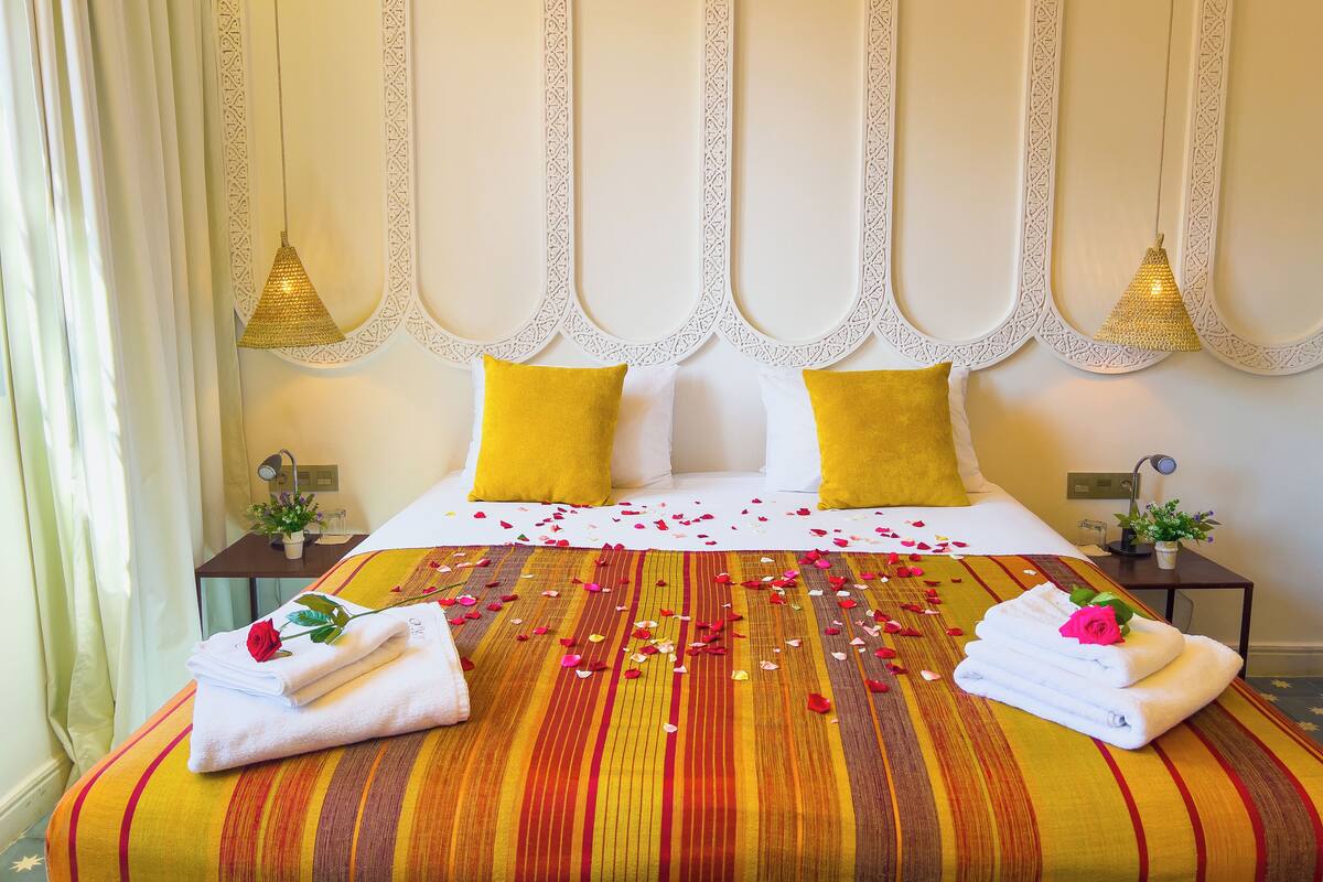 A hotel room with a large bed, two yellow pillows, a white headboard with decorative panels, a hanging light fixture, and a bedside table with a lamp and a small plant.
