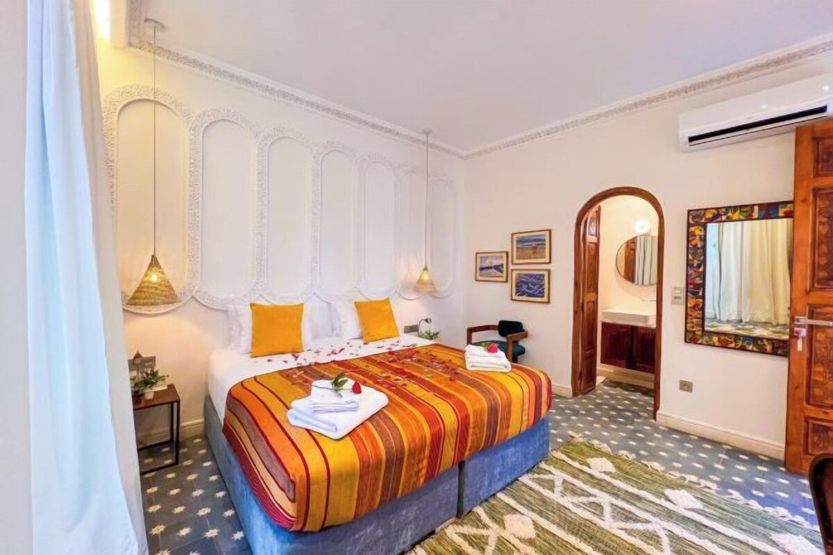 A hotel room with a large bed, a mirror, a small table, and a door leading to another room.