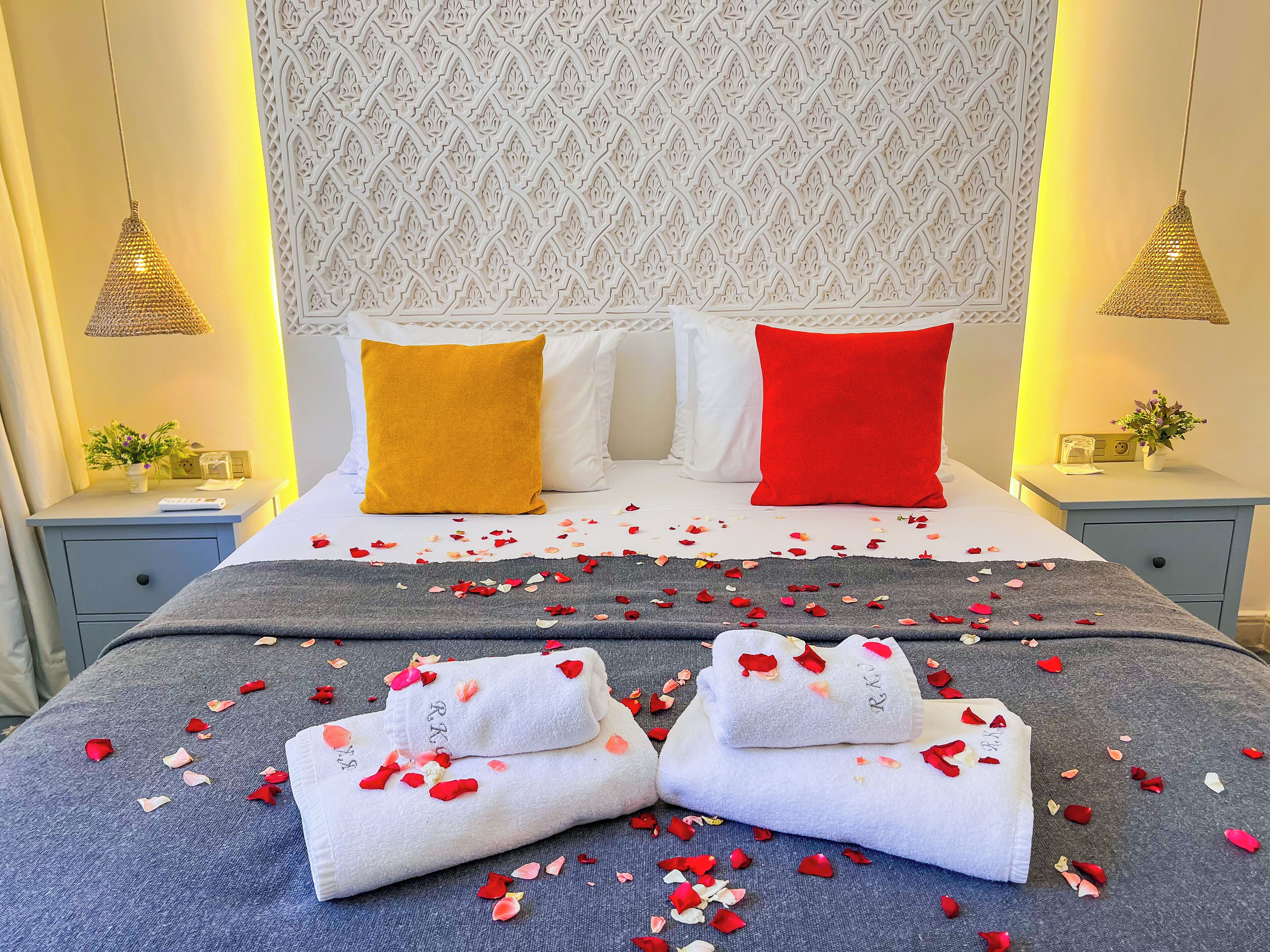 A neatly made bed with rose petals scattered across the bedspread, two white towels with initials, and a patterned wall behind.