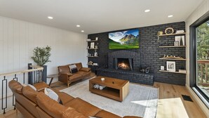A modern living room with a fireplace, leather sofas, and a large window offering a view of greenery. - Soaker Tubs + Decks | 5BR | Sleeps 12 (Lake Arrowhead)