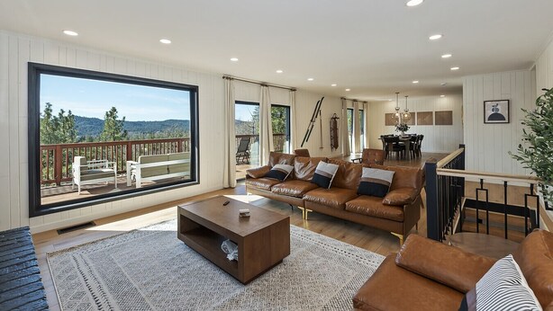 A spacious living room with a large window, a leather sofa, a coffee table, and a dining area. - Soaker Tubs + Decks | 5BR | Sleeps 12 (Lake Arrowhead)