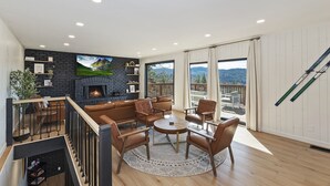 A modern living room with a fireplace, leather furniture, and a view of mountains through large windows. - Soaker Tubs + Decks | 5BR | Sleeps 12 (Lake Arrowhead)