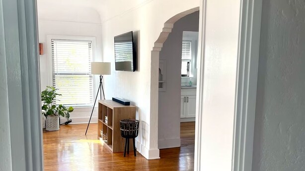 A spacious living room with hardwood flooring, a television mounted on the wall, and a potted plant.