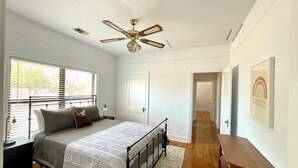 A bedroom with a bed, a ceiling fan, a window with blinds, a door, and a hallway leading to another room.