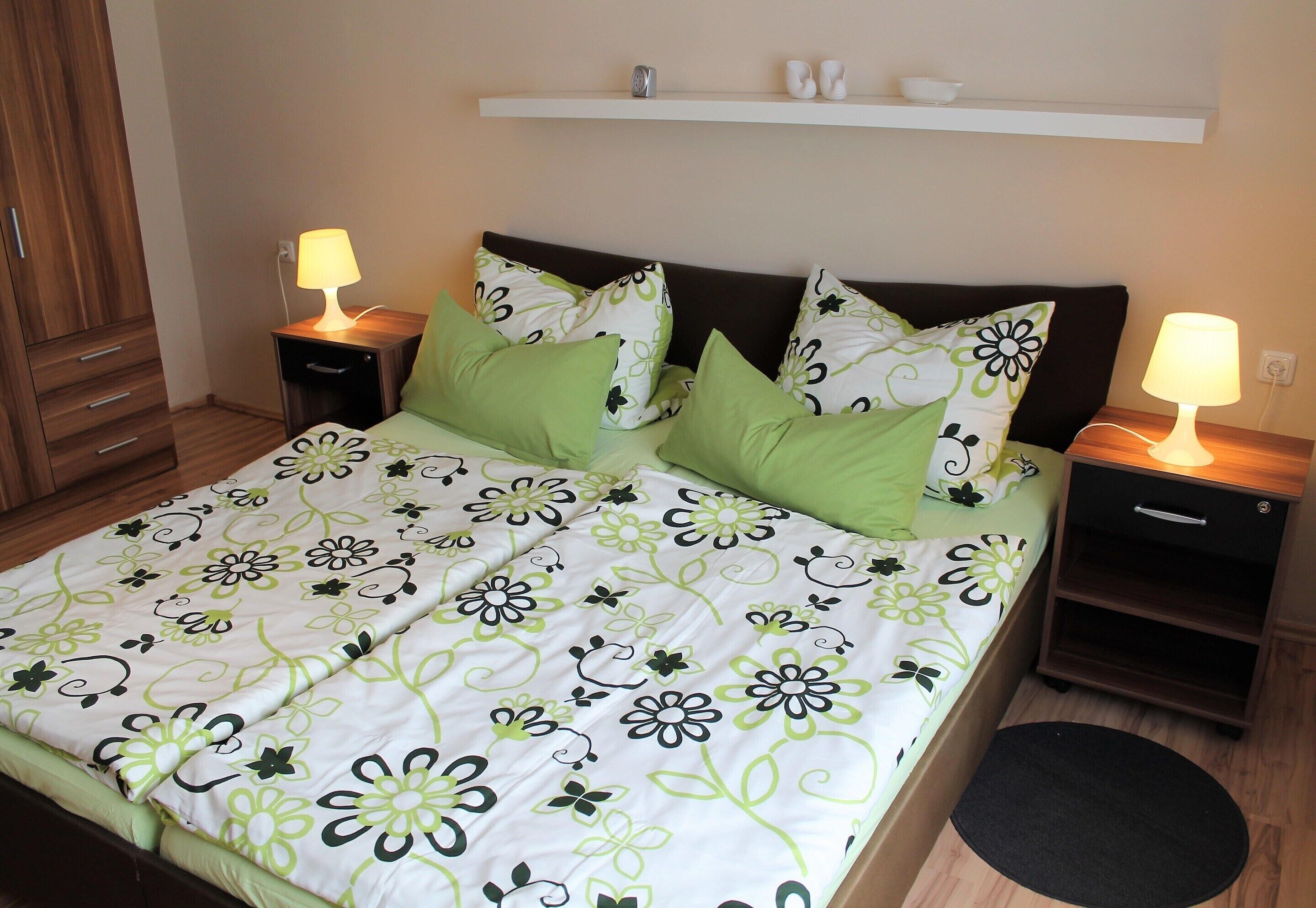 Ein ordentlich bezogenes Bett mit Blumenmuster, zwei Nachttische mit Lampen und ein kleines Regal darüber.
