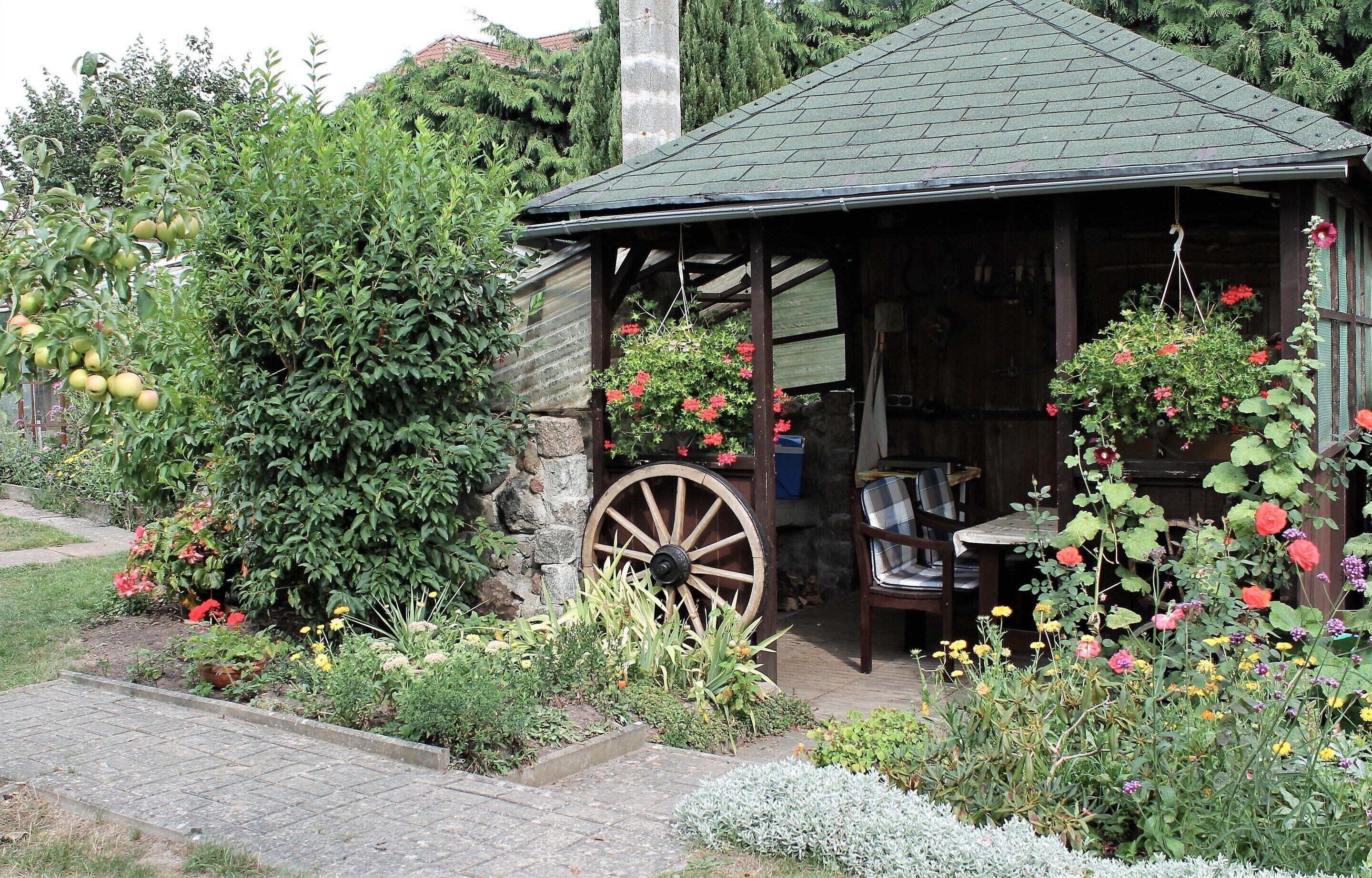 A rustic wooden structure with a thatched roof, a large wooden wheel, and a variety of flowering plants and trees.