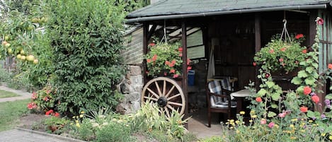 A rustic wooden structure with a thatched roof, a large wooden wheel, and a variety of flowering plants and trees.