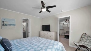 Un dormitorio con cama, ventilador de techo, televisor sobre una cómoda y una silla.